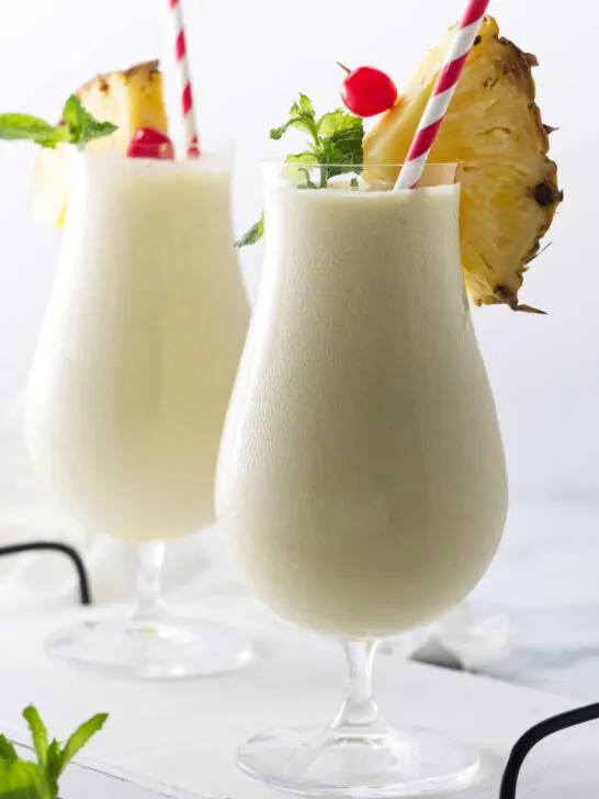 Two glasses of creamy non-alcohol beverage, with paper straws and garnishes of pineapple, cherry and mint.