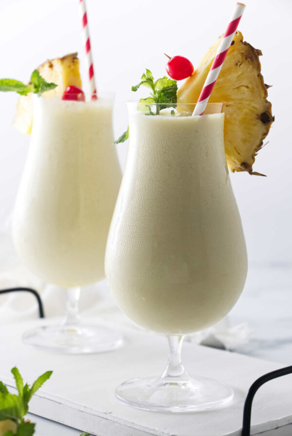 Two glasses of creamy non-alcohol beverage, with paper straws and garnishes of pineapple, cherry and mint.