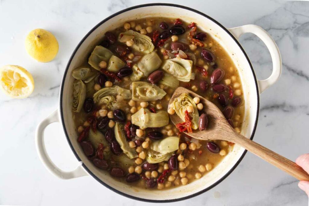 Pot with chicken, artichokes, olives and garbanzo beans ready to be cooked. Wooden spoon with sun-dried tomato, artichoke, olive and garbanzo beans.