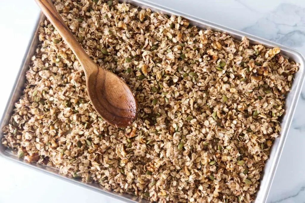 Half-sheet baking pan and a wooden spoon with mixed granola ready for baking.