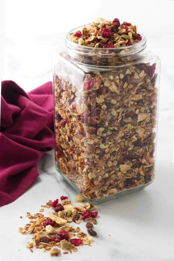 A large jar of granola with apples, cranberry granola spilled in front of the jar and a napkin in the background.