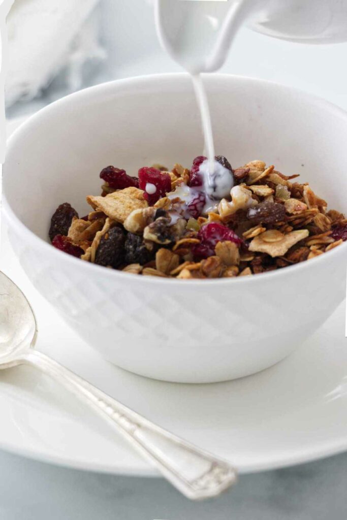 A dish with granola and milk being poured on top and a spoon alongside of the dish.