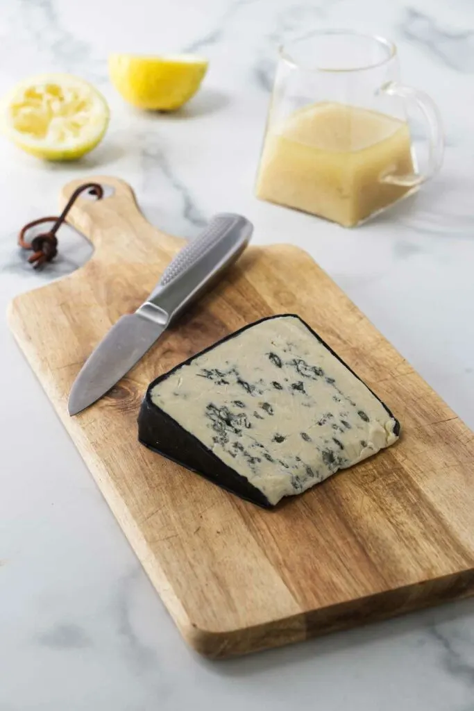 A wooden cutting board with a wedge of blue cheese and a paring knife. A small pitcher of lemon vinaigrette and lemon rinds in the background.