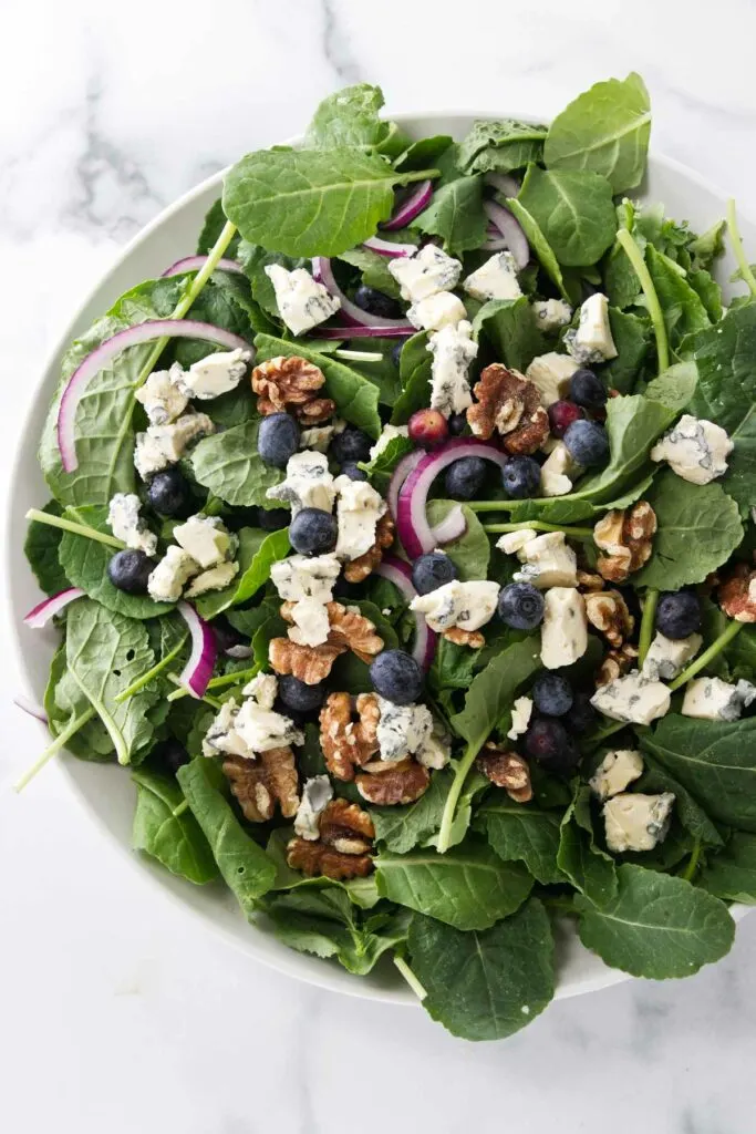 A large salad bowl filled with baby kale, blueberries, red onion slices and roasted walnuts.