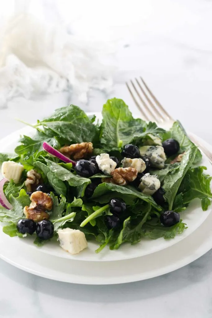 A serving of baby kale and blueberry salad on a salad plate with a fork and a cloth napkin in the background.