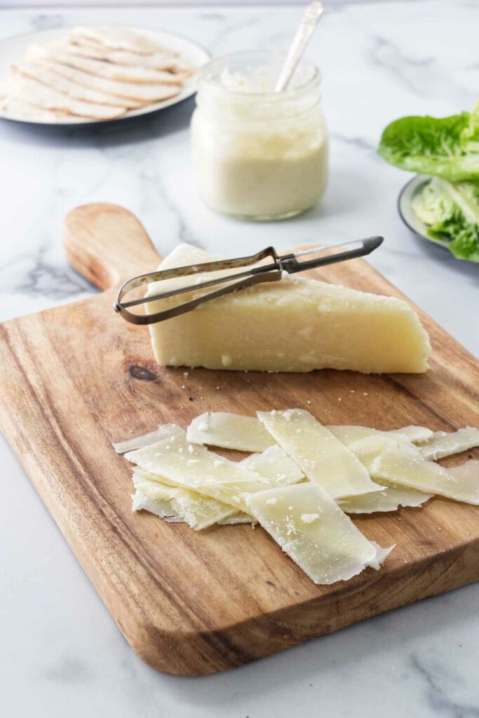 A wedge of parmesan cheese with a vegetable peeler and shavings o the cheese on a wooden cutting board. A plate with lettuce, a jar of Caesar dressing and a plate of sliced chicken in the background.