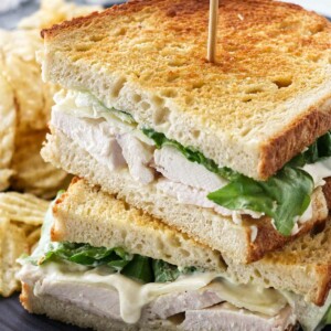 Two halves of a chicken sandwich stacked together with a wooden pick plus potato chips on a plate.