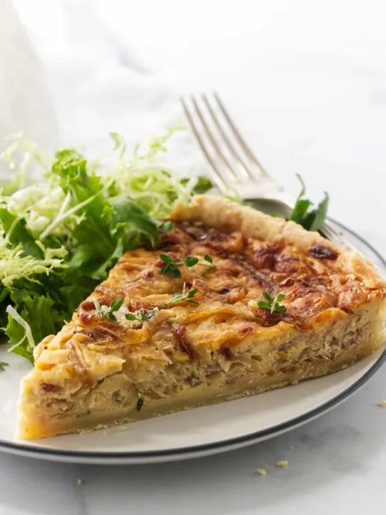 A serving of French onion tart on a plate with a fork. Green salad in the background.