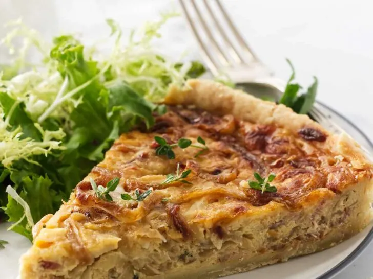 A serving of French onion tart on a plate with a fork. Green salad in the background.