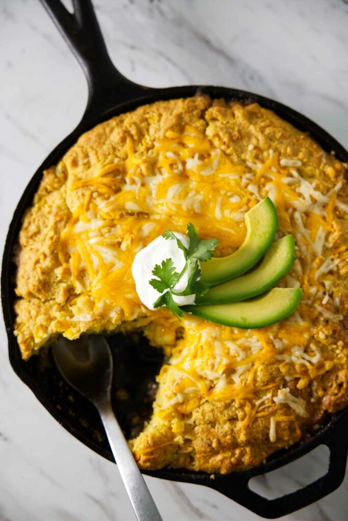 A green chili tamale pie in a skillet with sour cream and avocado slices on top.