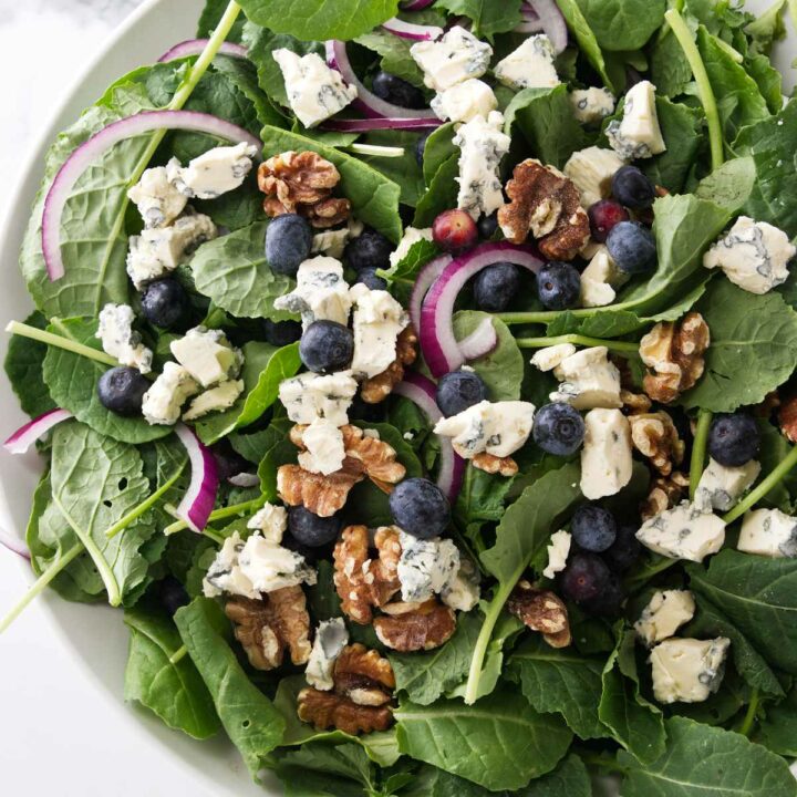 A large bowl of salad greens with blueberries, red onion slices roasted walnuts and blue cheese.