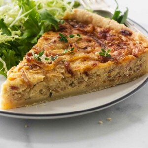 A serving of French onion tart on a plate with a fork. Green salad in the background.