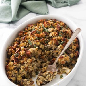 A dish of stuffing with a serving spoon, a kitchen towel in the background.