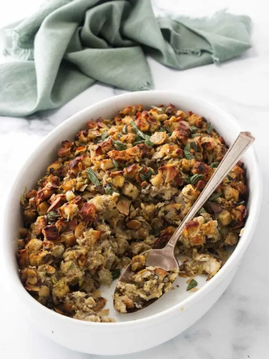 A dish of stuffing with a serving spoon, a kitchen towel in the background.
