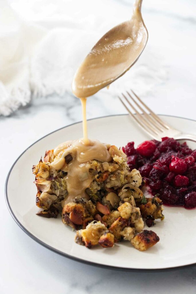 A serving of stuffing on a plate with gravy being drizzled on the stuffing from a spoon. Cranberries and a fork next to the stuffing.