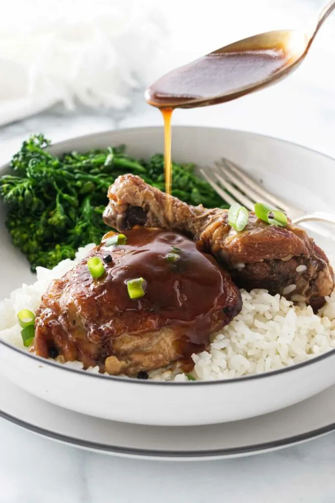 Spooning sauce over a serving of chicken on a bed of rice with a side of broccolini.