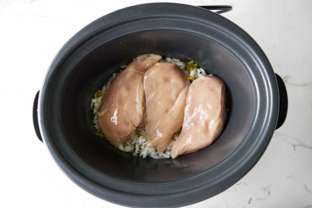 A crockpot with chicken and diced onions inside.