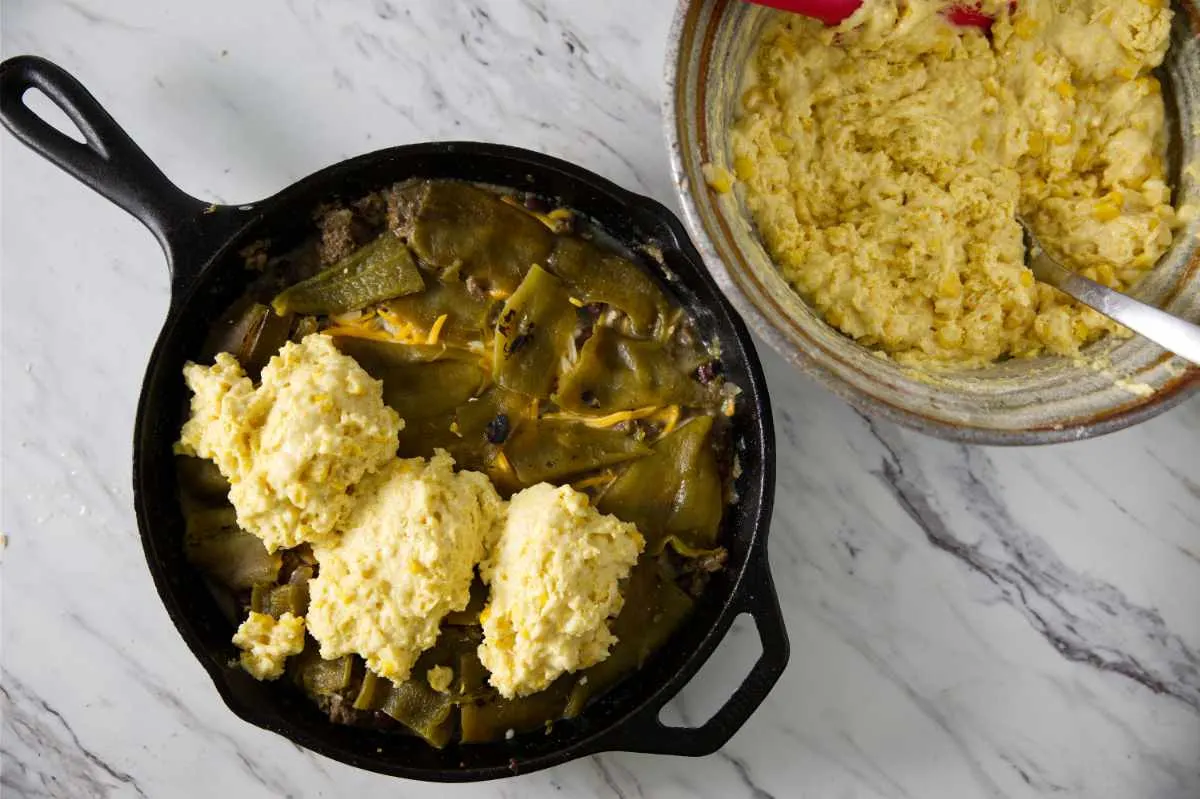 Adding cornbread batter to the top of tamale filling in a skillet.