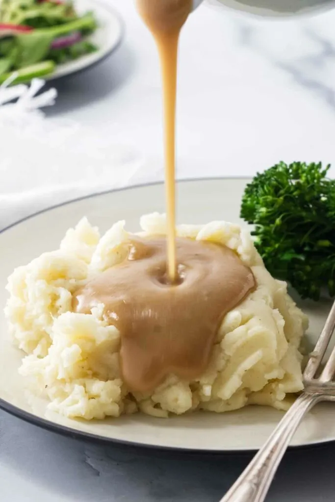 A serving plate with mashed potatoes and gravy being poured on the potatoes. A napkin and side salad in the background.