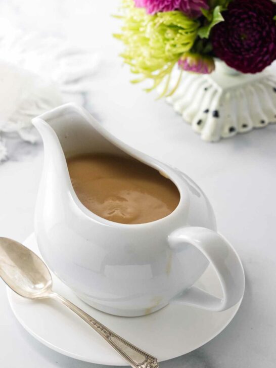 A pitcher of turkey gravy on a saucer with a spoon. A napkin and flowers in the background.