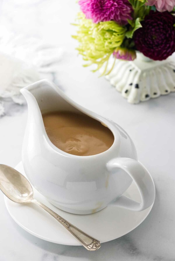 A pitcher of turkey gravy on a saucer with a spoon. A napkin and flowers in the background.