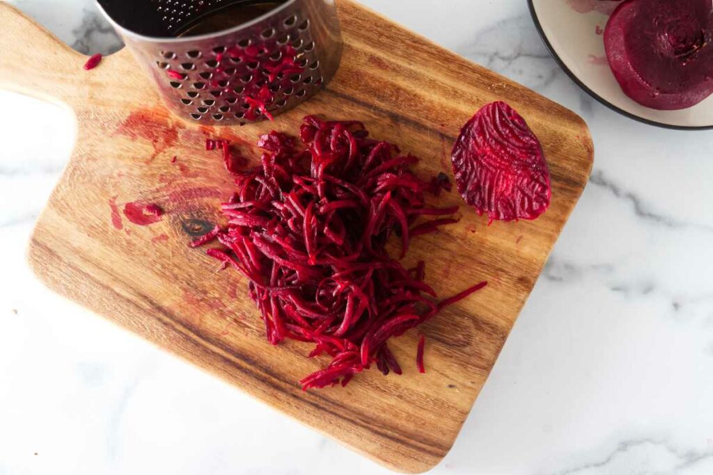 Grated beets on a cutting board with a box grater. A plate with a whole beet in the upper right corner.