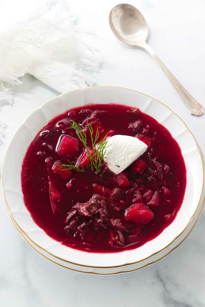 A soup bowl of borscht garnished with sour cream and a sprig of dill. A soup spoon and napkin in the background.