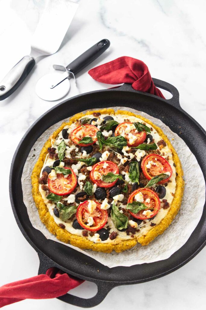 Baked polenta crust pizza on a parchment paper-lined pizza pan. Pizza utensils in the upper left corner.
