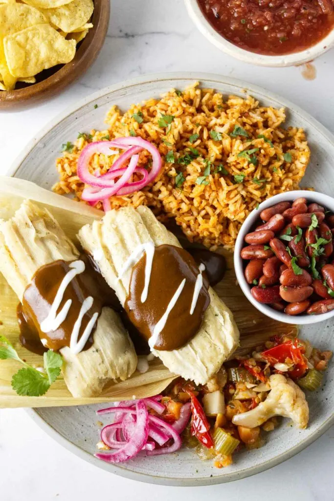 A dinner plate filled with a serving of tamales, Mexican rice, pinto beans, and pickled veggies. Corn chips and salsa are on the side.