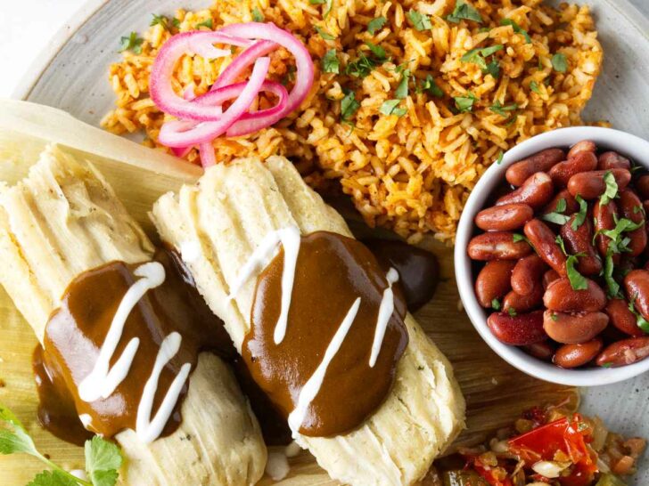 A dinner plate filled with a serving of tamales, Mexican rice, pinto beans, and pickled veggies. Corn chips and salsa are on the side.