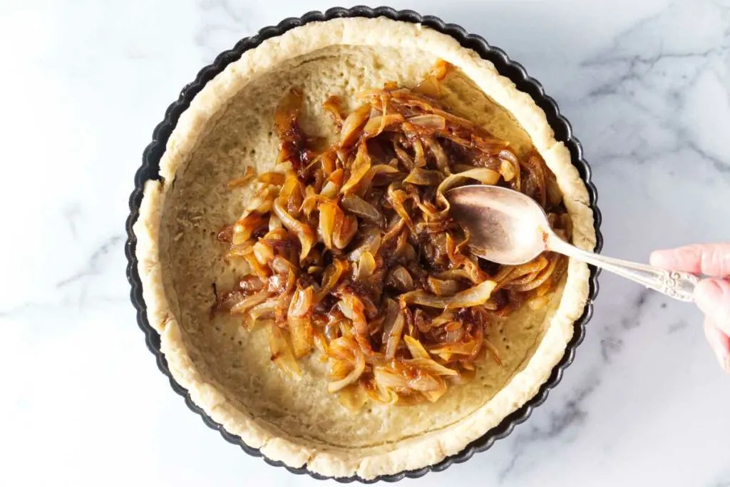 A serving spoon adding caramelized onions to a par-baked quiche crust.