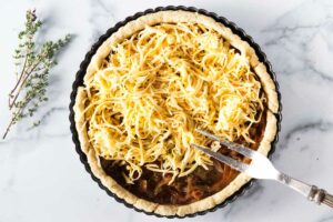 A portion of shredded cheese being spread on an unbaked onion quiche with a serving fork.
