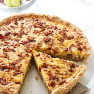 Cheese and onion quiche on a serving plate with a pie server lifting up a slice for serving. A plate of green and red grapes in the background.