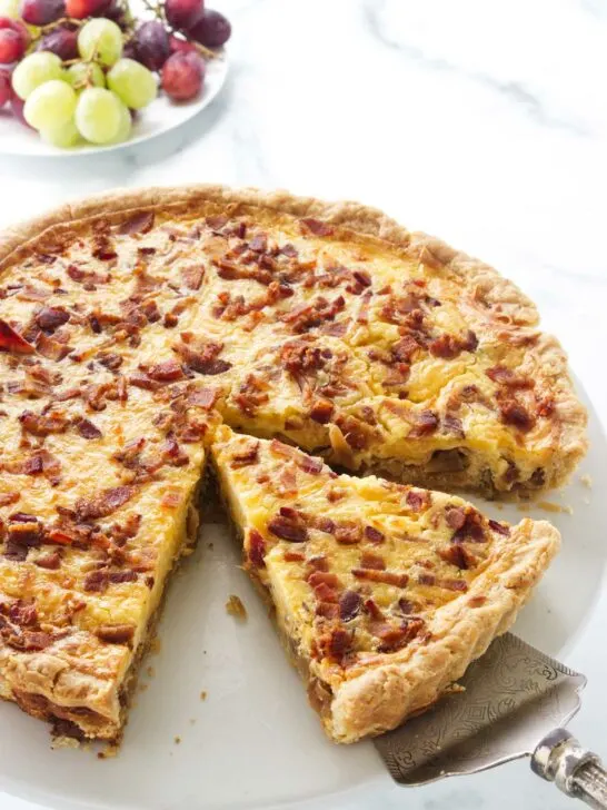 Cheese and onion quiche on a serving plate with a pie server lifting up a slice for serving. A plate of green and red grapes in the background.