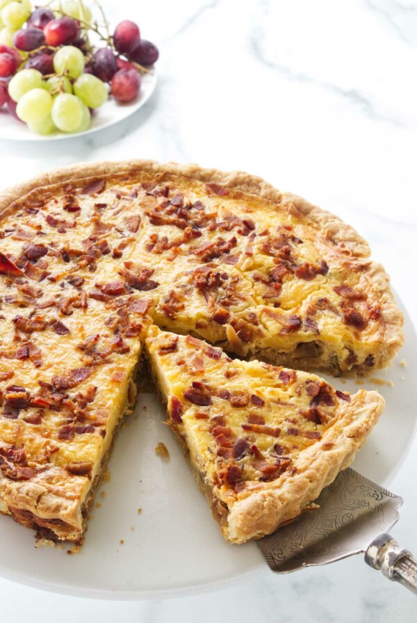 Cheese and onion quiche on a serving plate with a pie server lifting up a slice for serving. A plate of green and red grapes in the background.