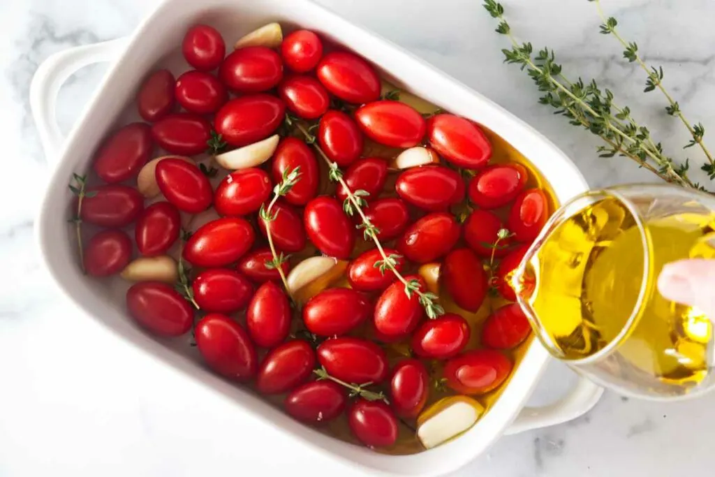 Pouring olive oil in a dish with tomatoes, garlic and thyme sprigs.