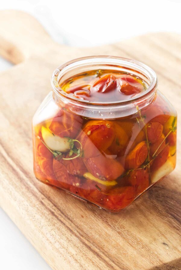 Small jar of tomatoes, garlic and thyme sprigs covered in olive oil and sitting on a cutting board.