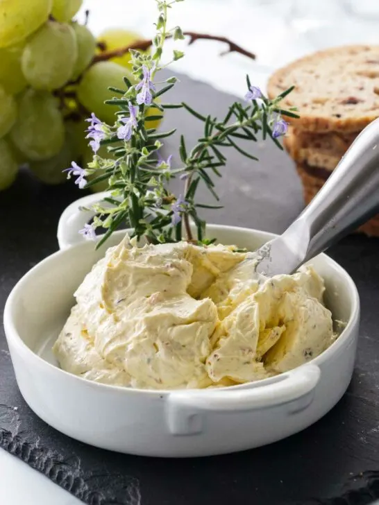 A dish of butter mixture with an appetizer knife garnished with fresh rosemary sprigs. Grapes and crackers in the background.