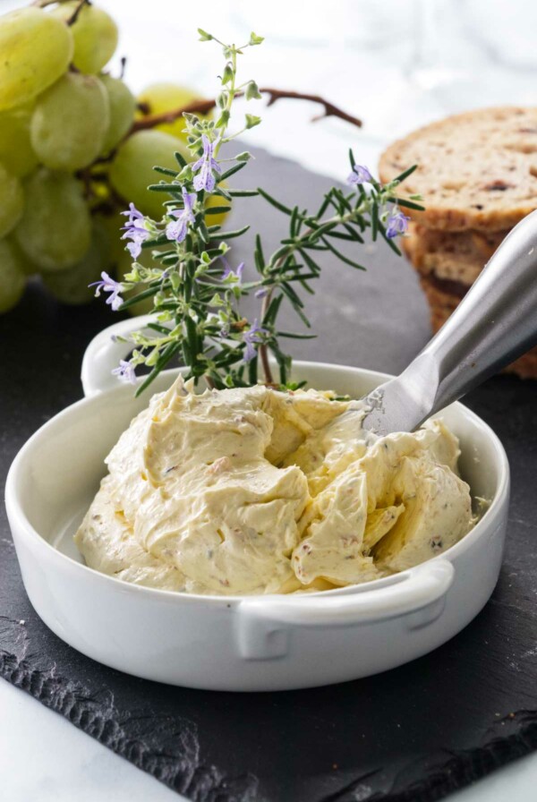 A dish of butter mixture with an appetizer knife garnished with fresh rosemary sprigs. Grapes and crackers in the background.