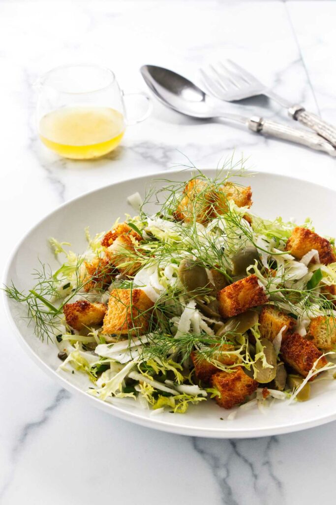 A bowl of salad with croutons, olives, fennel, fennel fronds and lettuce. Salad dressing and serving utensils in the background.