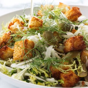 Pouring a salad dressing over a salad of fennel, croutons, lettuce, green olives and capers.