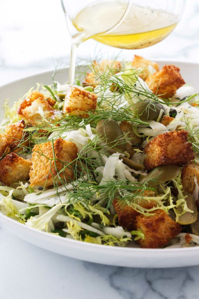 Pouring a salad dressing over a salad of fennel, croutons, lettuce, green olives and capers.