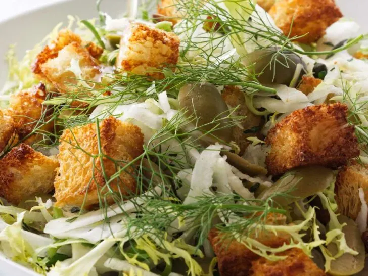 Pouring a salad dressing over a salad of fennel, croutons, lettuce, green olives and capers.