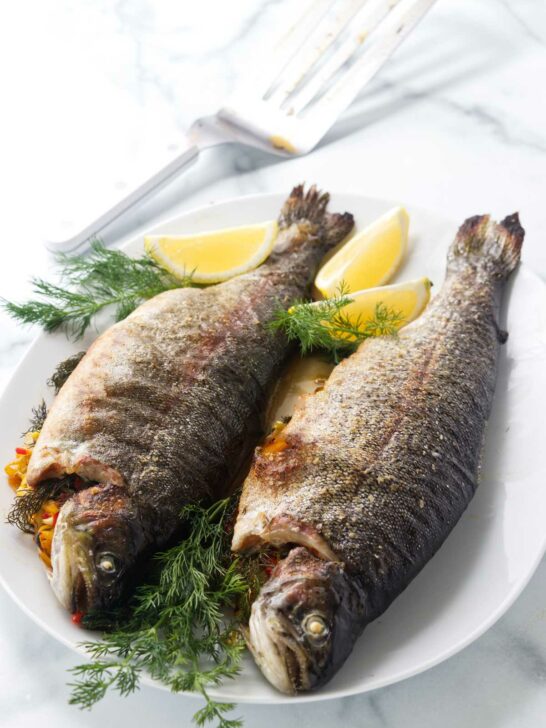 Two baked stuffed trout on a platter with lemon wedges and fresh dill.