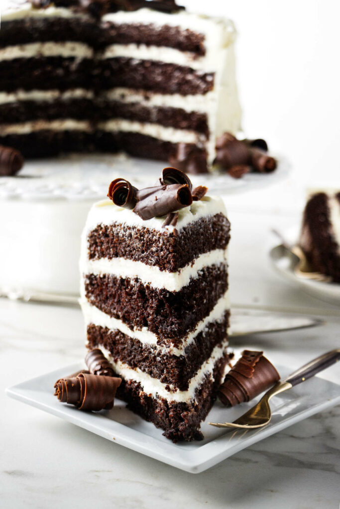 A dark chocolate cake with cream cheese icing on a platter with a slice of cake in the foreground.