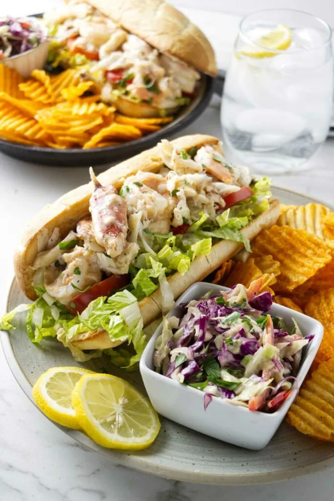 Two seafood po boys on plates with coleslaw and potato chips.