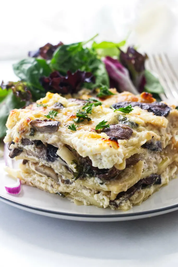 A serving of mushroom lasagna on a plate with side salad and fork.