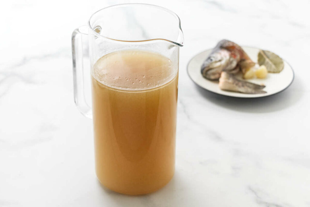 A pitcher of Homemade Fish Broth, and saucer of fish head, tail, garlic and bay leaves in the background.