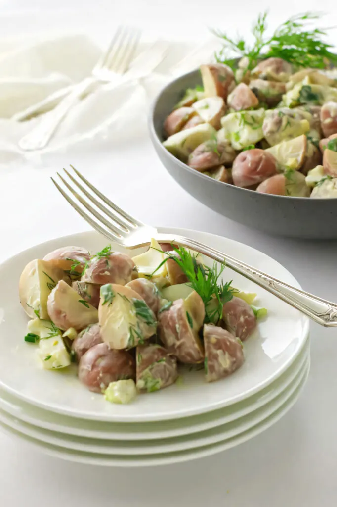 A bowl and plate filled with potato salad made with red potatoes and yogurt dressing.
