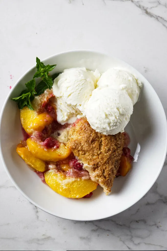 A serving of peach raspberry cobbler in a bowl with ice cream.